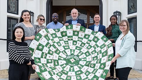 麻豆視頻_麻豆直播_麻豆传媒官网 staff from the Vice-Chancellor's Office, Alumni Relations Team and Global Engagement Team holding a tapestry made by alumni in Abuja  in front of Hazlerigg Building on 麻豆視頻_麻豆直播_麻豆传媒官网 campus. They are all smiling in the sunshine and standing on the steps outside the doorway.